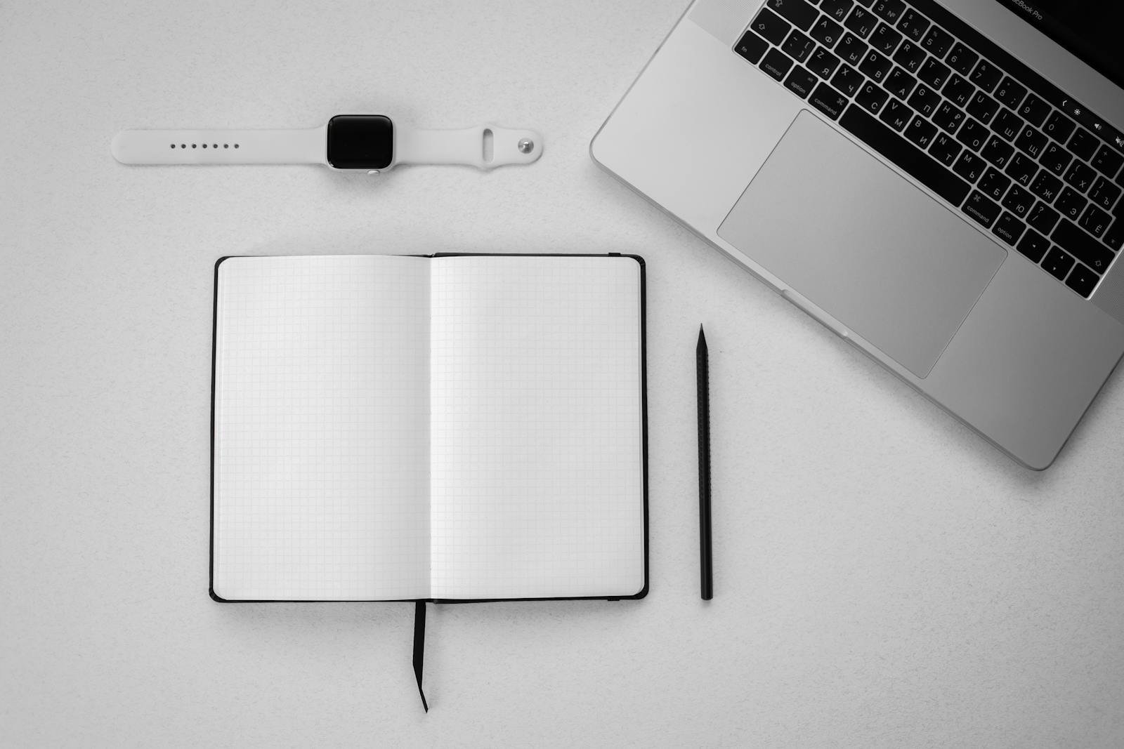Top-down view of a minimalist workspace featuring a laptop, blank notebook, smartwatch, and pencil.